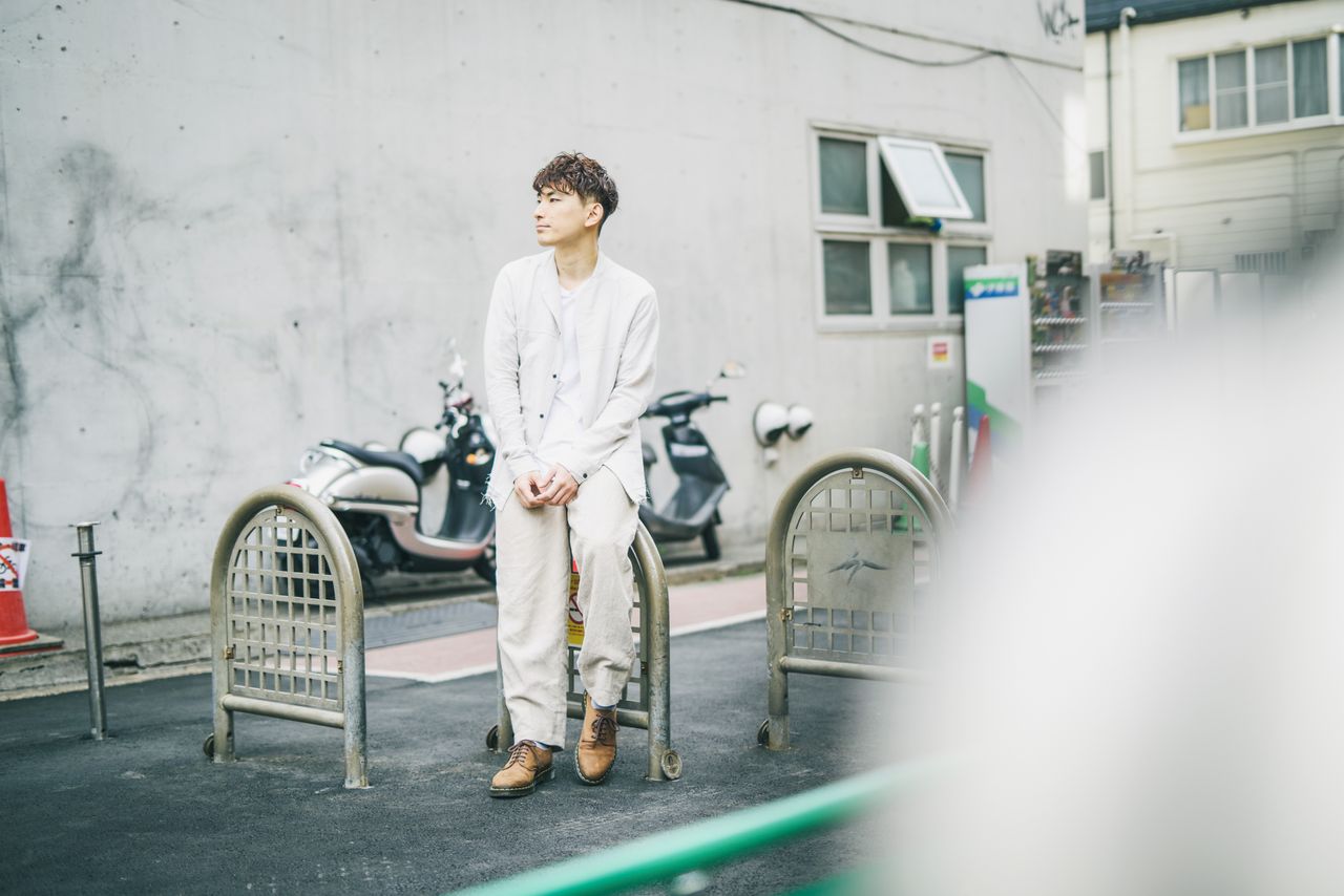 Asian man wearing white suit sitting on a sidewalk with motorbikes in the background.